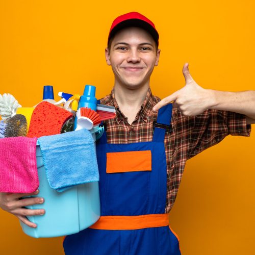 pleased young cleaning guy wearing uniform and cap holding and points at bucket of cleaning tools isolated on orange background