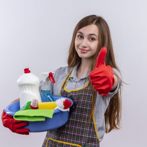 young beautiful girl in apron and rubber gloves holding basin with cleaning tools smiling showing thumbs up standing over white background